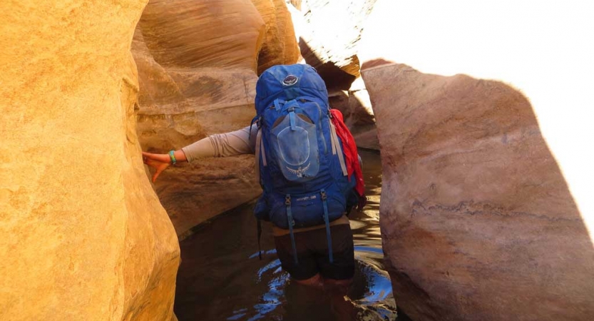 A person wearing a backpack wades through thigh-deep water between tight canyon walls